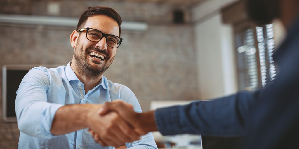 A smiling man with glasses shaking hands with another person in a brightly lit office setting.