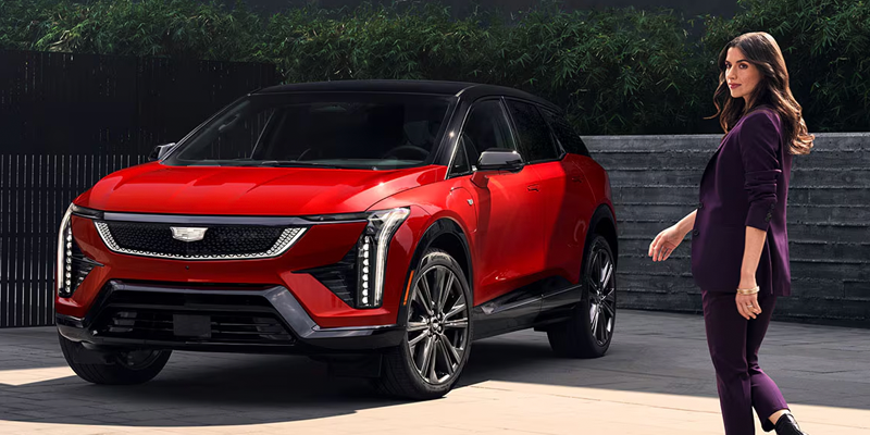 Front-three-quarter view of a red Cadillac OPTIQ electric SUV with a black roof, parked outdoors as a woman in a purple suit walks by in the foreground.