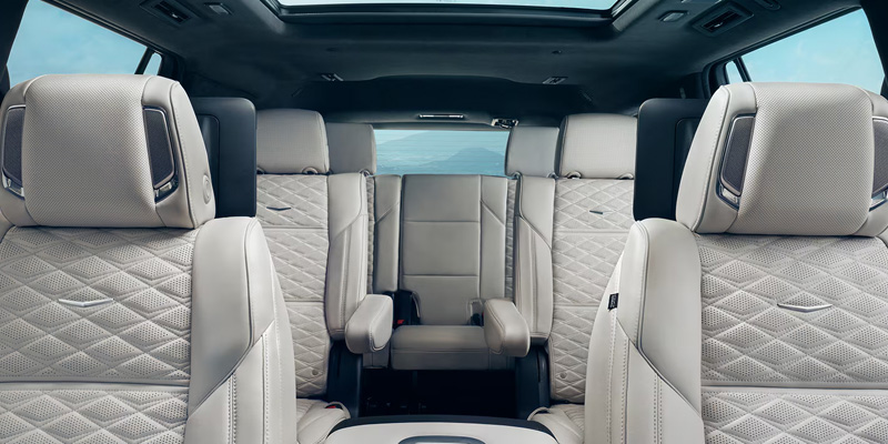 Interior view looking back at the three rows of light gray quilted leather seats in a Cadillac Escalade, featuring front headrest speakers, second-row captain's chairs with rear screens, and a sunroof.
